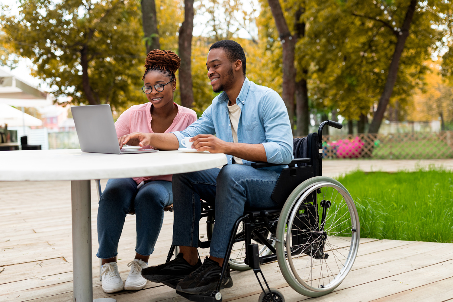 A woman and man in a wheelchair on their laptop at a table outside.