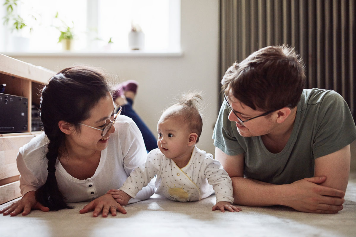 A mom, baby and dad are on the floor doing tummy time together. Both parents are looking and smiling at the baby.
