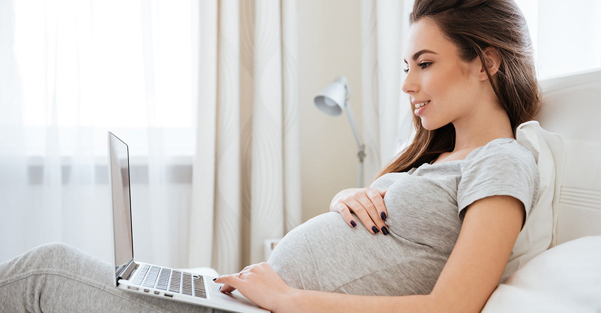 pregnant woman smiling, looking at laptop in bed