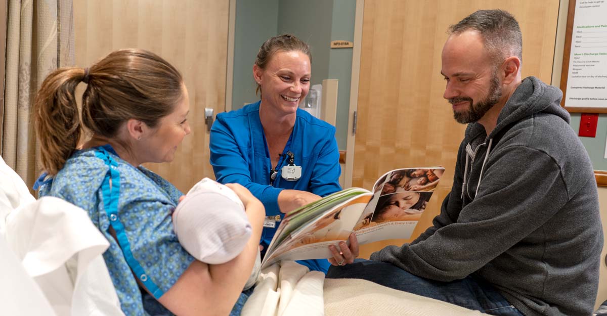 Nurse stitting bedside with new mom holding baby and dad showing them the Understanding Postpartum health and Baby Care book